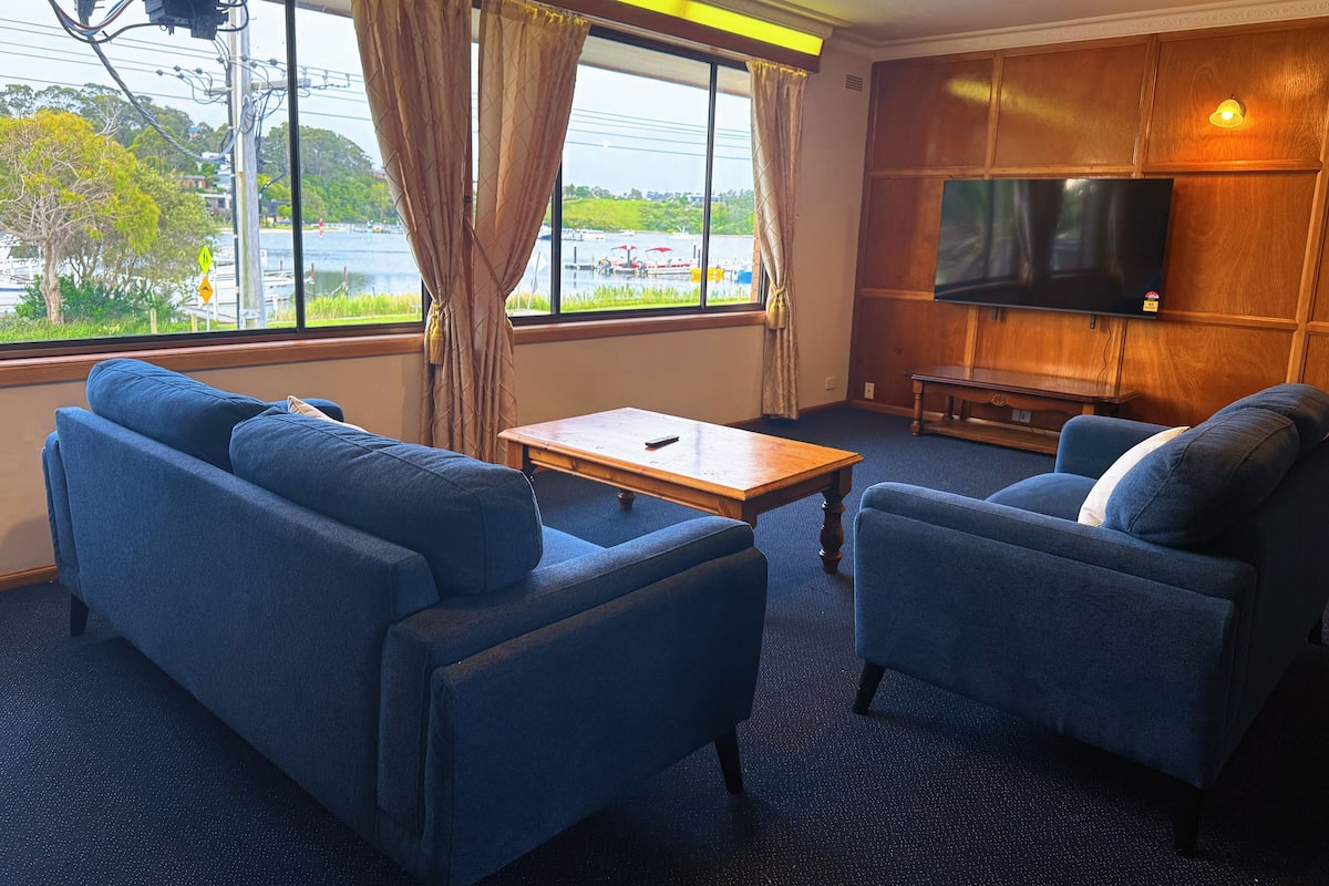 A hotel room with a large window, a sofa, a chair, a small table, and a flat-screen TV mounted on the wall.
