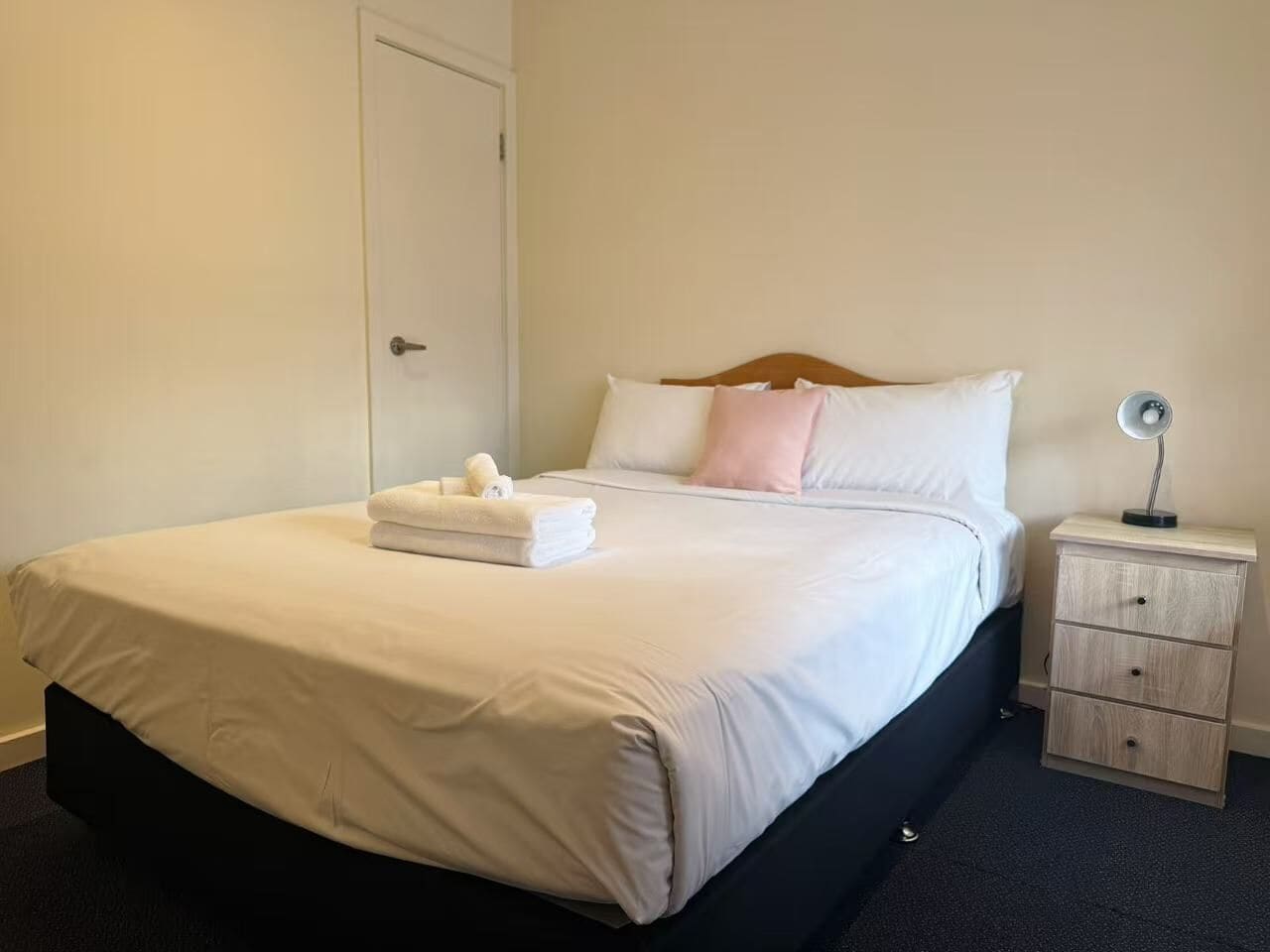 Un lit bien fait, avec du linge de lit blanc et un coussin rose, une table de chevet avec une lampe, et une porte visible au fond.
