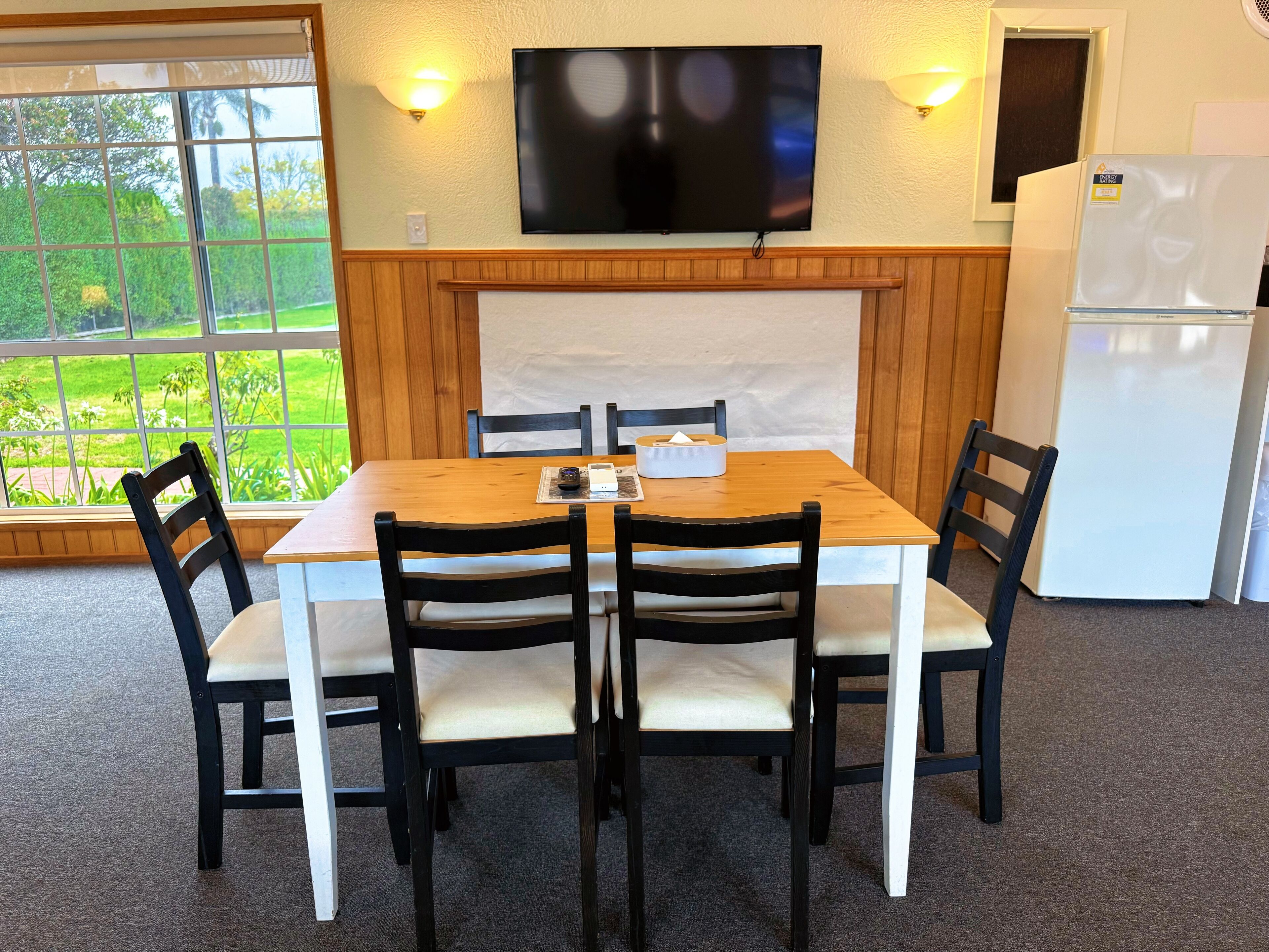 Un comedor con una mesa de madera y cuatro sillas, un televisor de pantalla plana montado arriba y un refrigerador a la derecha.