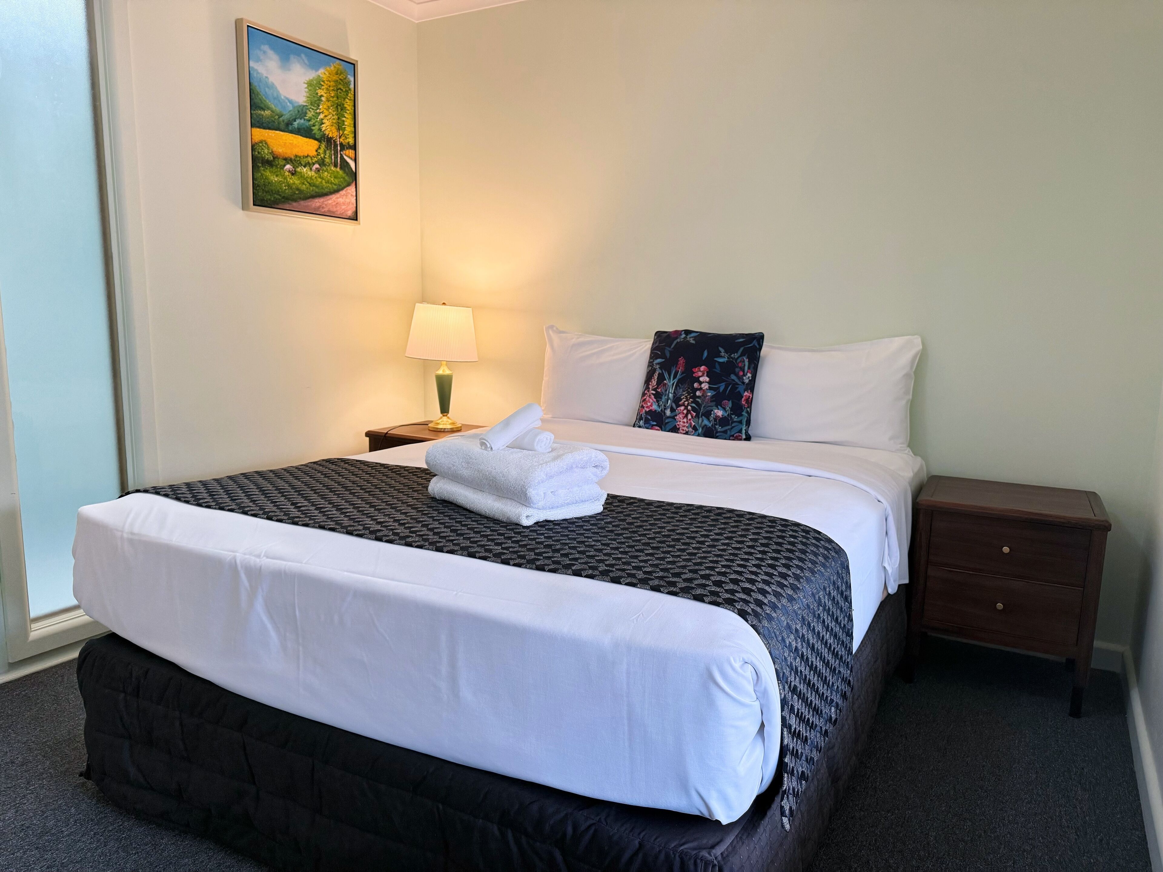 Una cama bien hecha con sábanas blancas, una toalla enrollada, una mesita de noche con una lámpara, un cojín floral y un cuadro de un paisaje en la pared.