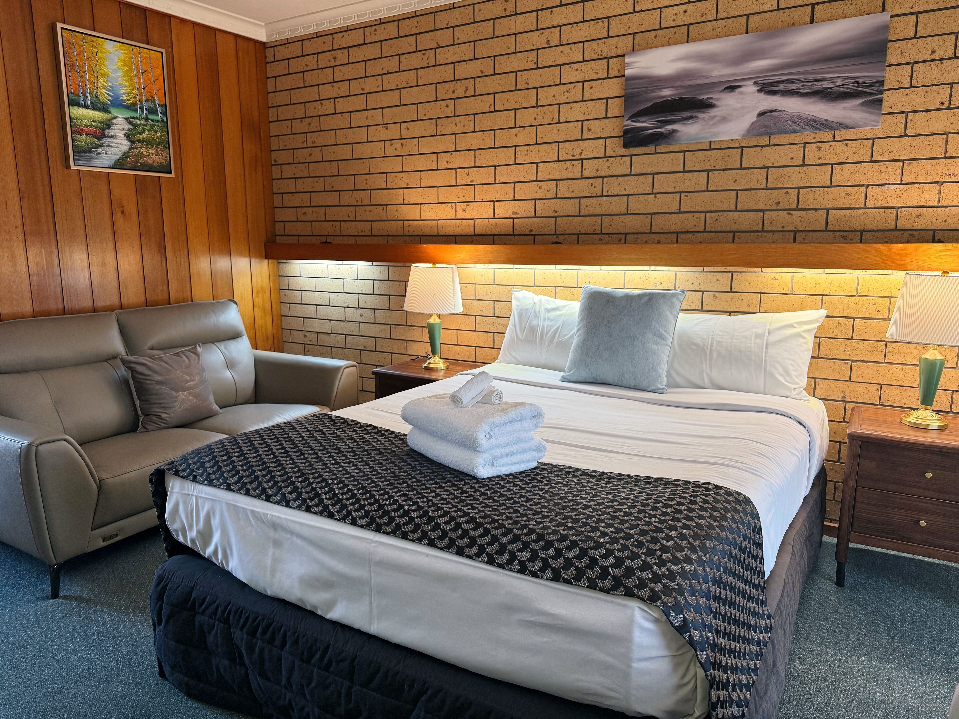 A hotel room with a brick wall, a bed with white and patterned bedding, a brown leather sofa, and two bedside tables with lamps.