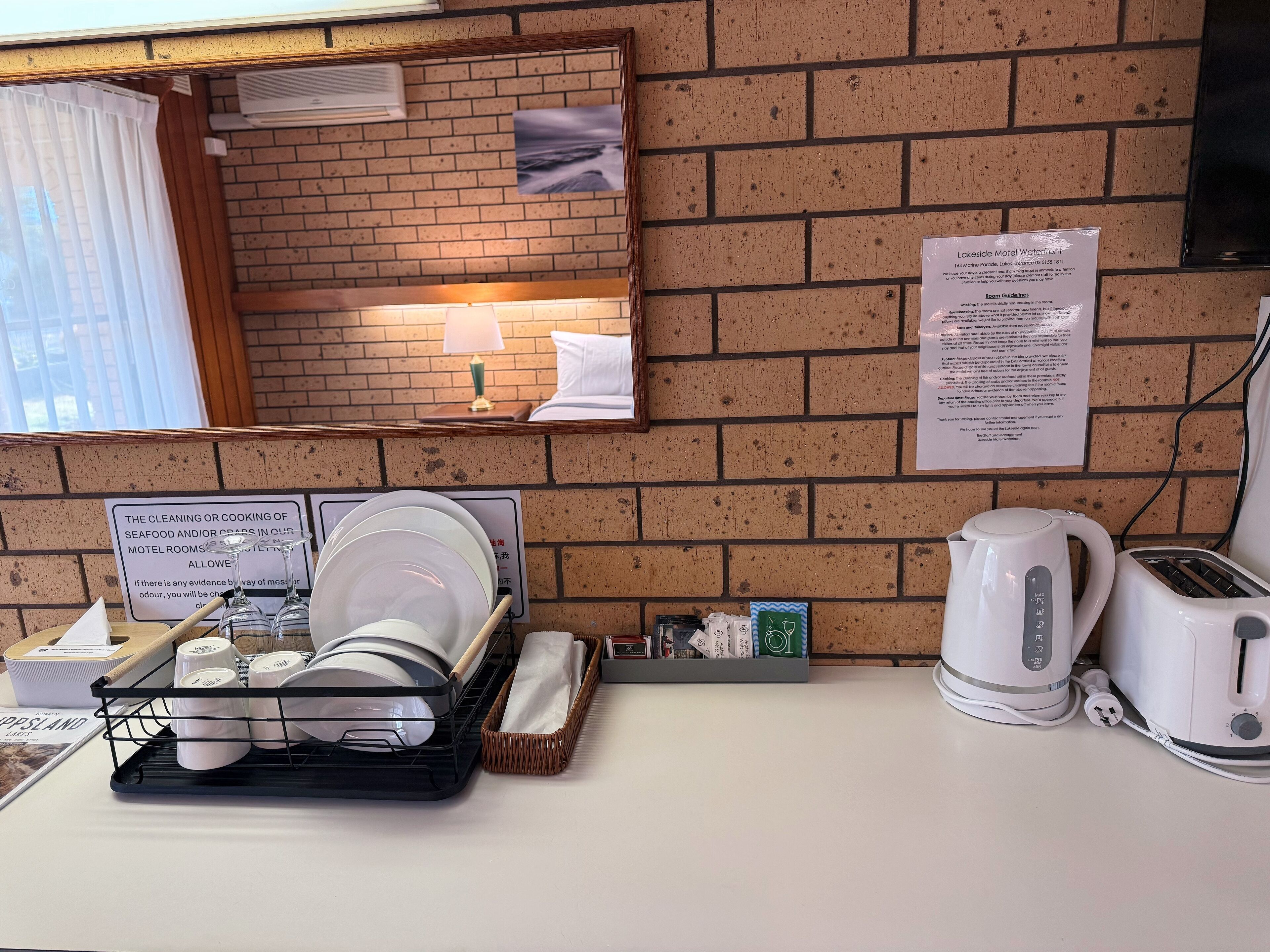 A hotel room with a brick wall, a mirror, a lamp, a sign with room guidelines, a dish rack with plates and glasses, a kettle, and a toaster.