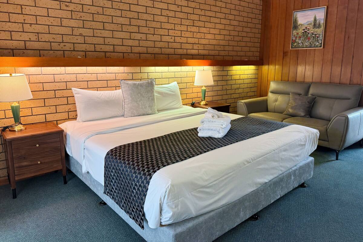 A hotel room with a brick wall, a bed with white linens, two bedside tables with lamps, a grey sofa, and a painting on the wall.