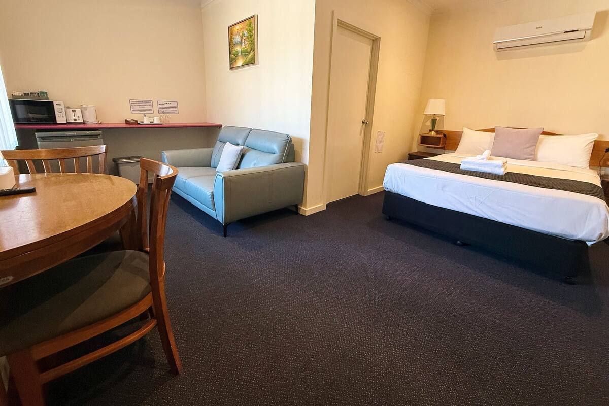A hotel room with a bed, a sofa, a dining table, and a kitchen area with appliances.
