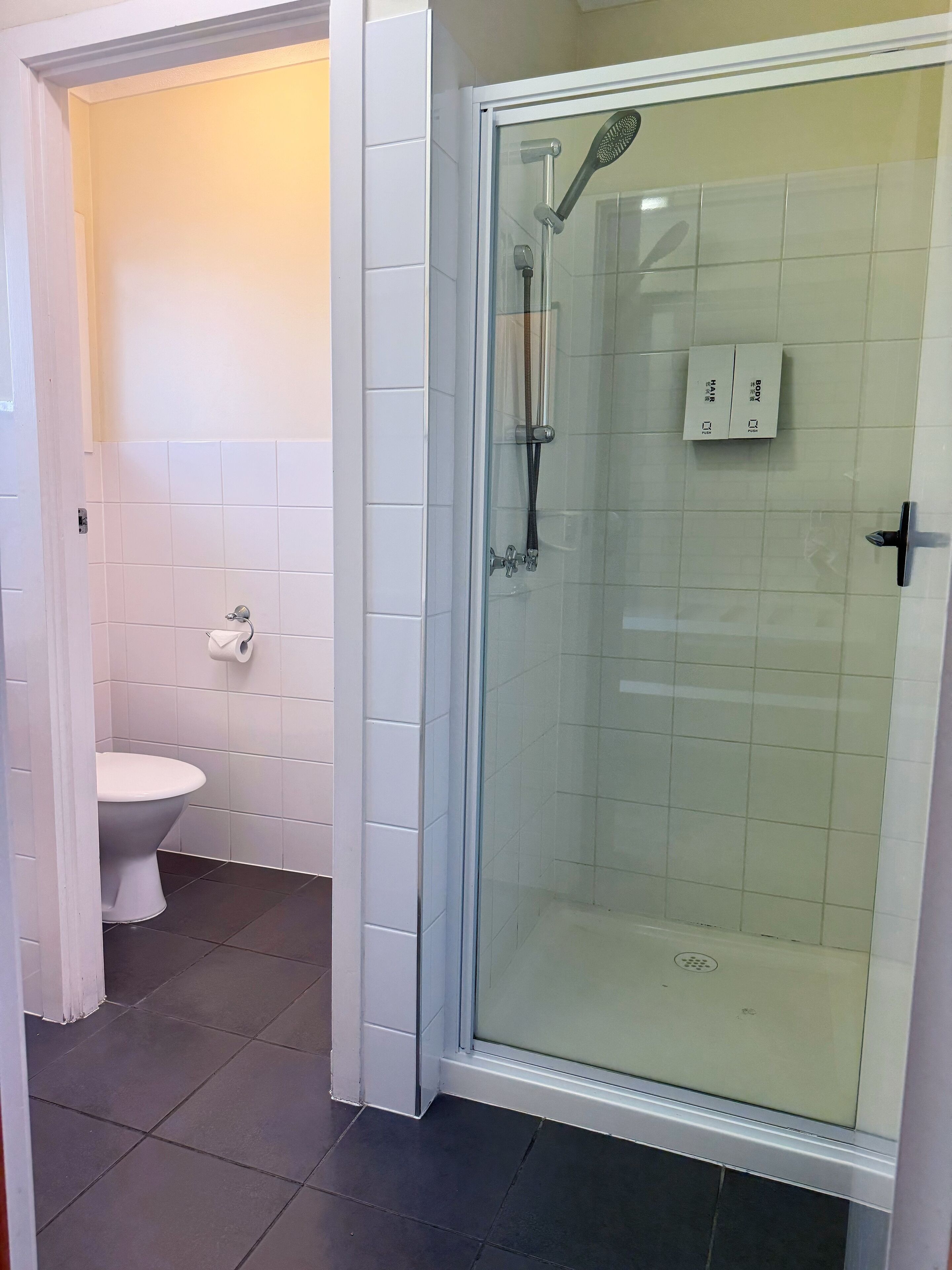 Une salle de bain avec des toilettes, une douche à parois de verre et des murs recouverts de carreaux blancs.