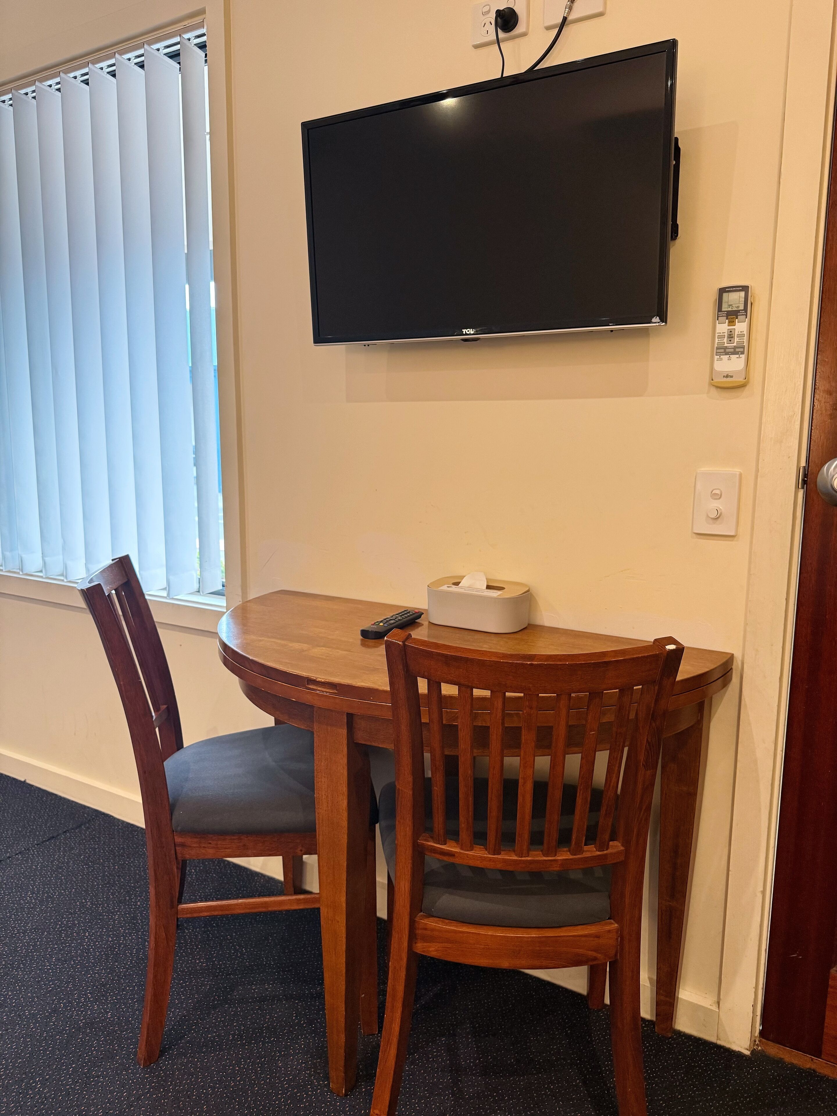 A room with a wooden dining table, two chairs, a flat-screen TV mounted on the wall, a window with blinds, and a thermostat.