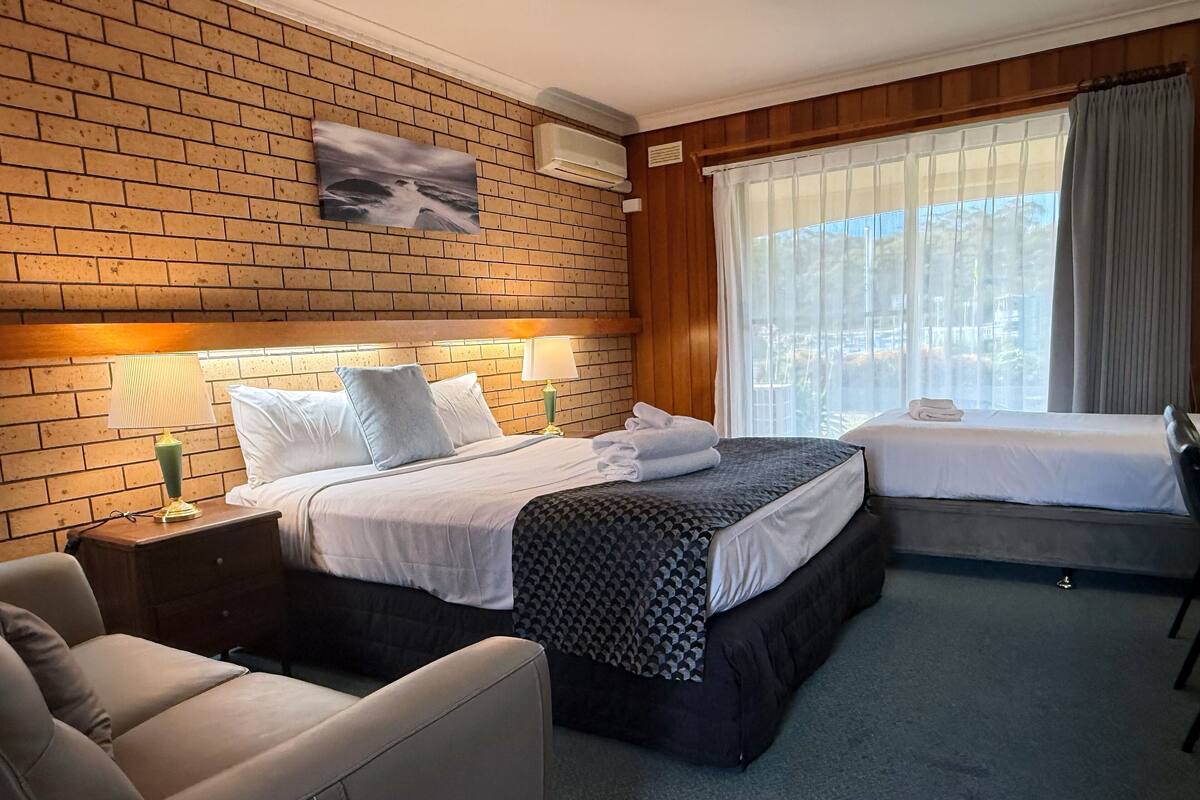 A hotel room with a brick wall, a bed with a patterned bedspread, a nightstand with a lamp, a chair, and a window with curtains.