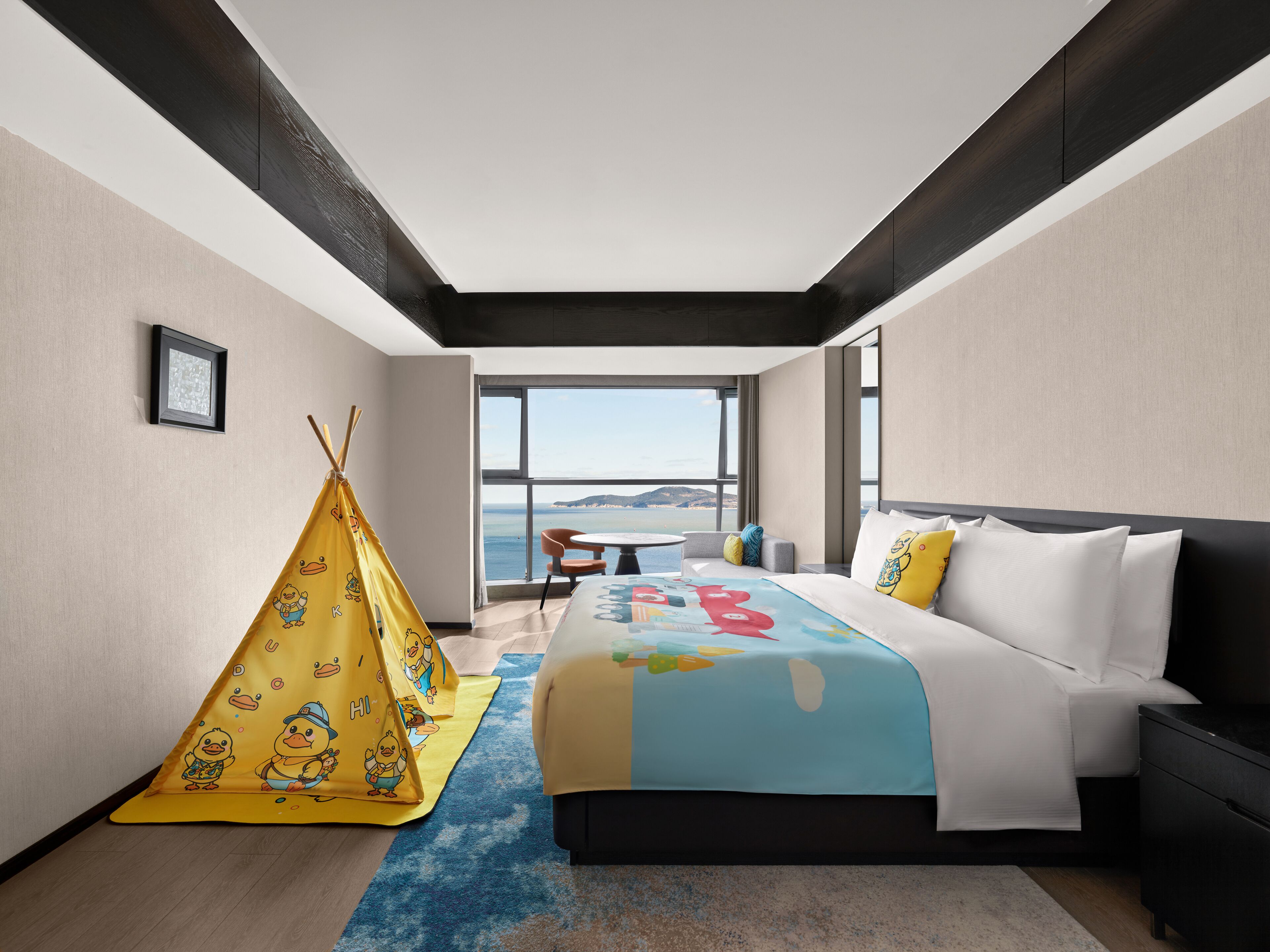 Habitación de hotel moderna con una cama grande, vista al mar, un tipi amarillo y una pequeña mesa con sillas.