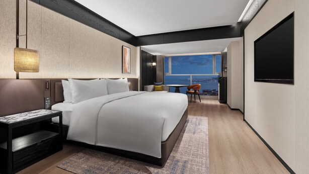 A modern hotel room with a large bed, a desk, a chair, a TV, and a view of the city.