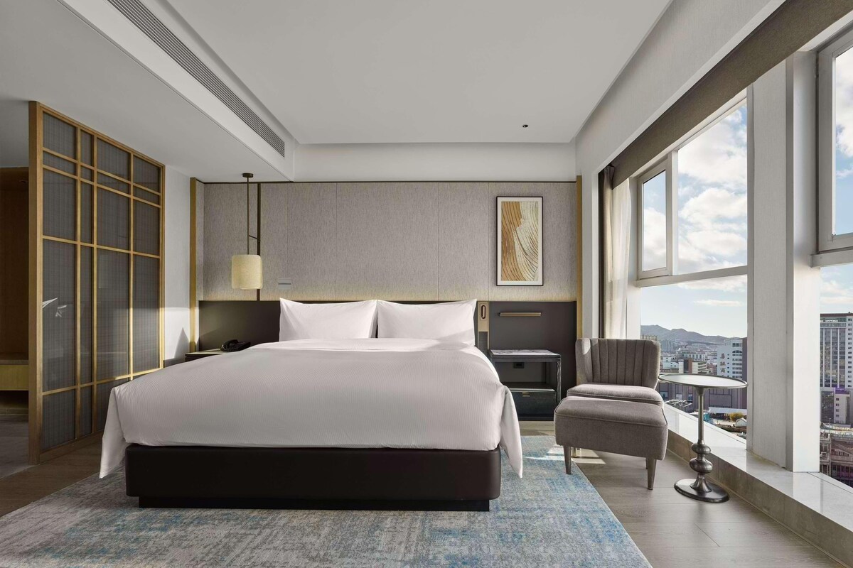 Een moderne hotelkamer met een groot bed, een zithoek, uitzicht op de stad en een groot raam.