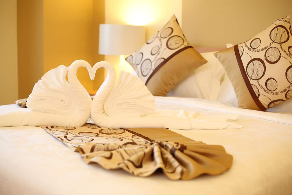 A neatly made bed with a heart-shaped towel arrangement, patterned pillows, and a folded towel on the bed.