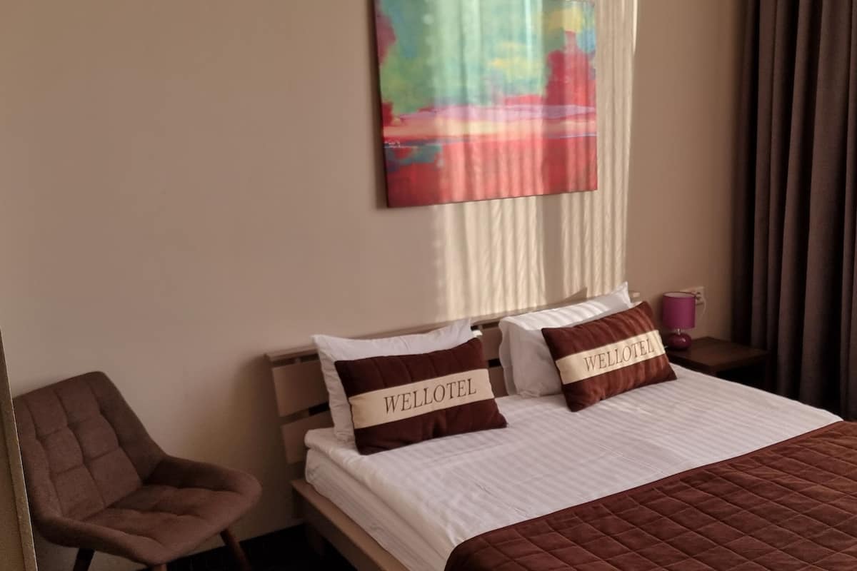 Ein Hotelzimmer mit einem Bett, zwei Kissen mit dem Schriftzug 'WELLLOTEL', einem Sessel, einem Nachttisch mit einer violetten Lampe und einem Gemälde an der Wand.