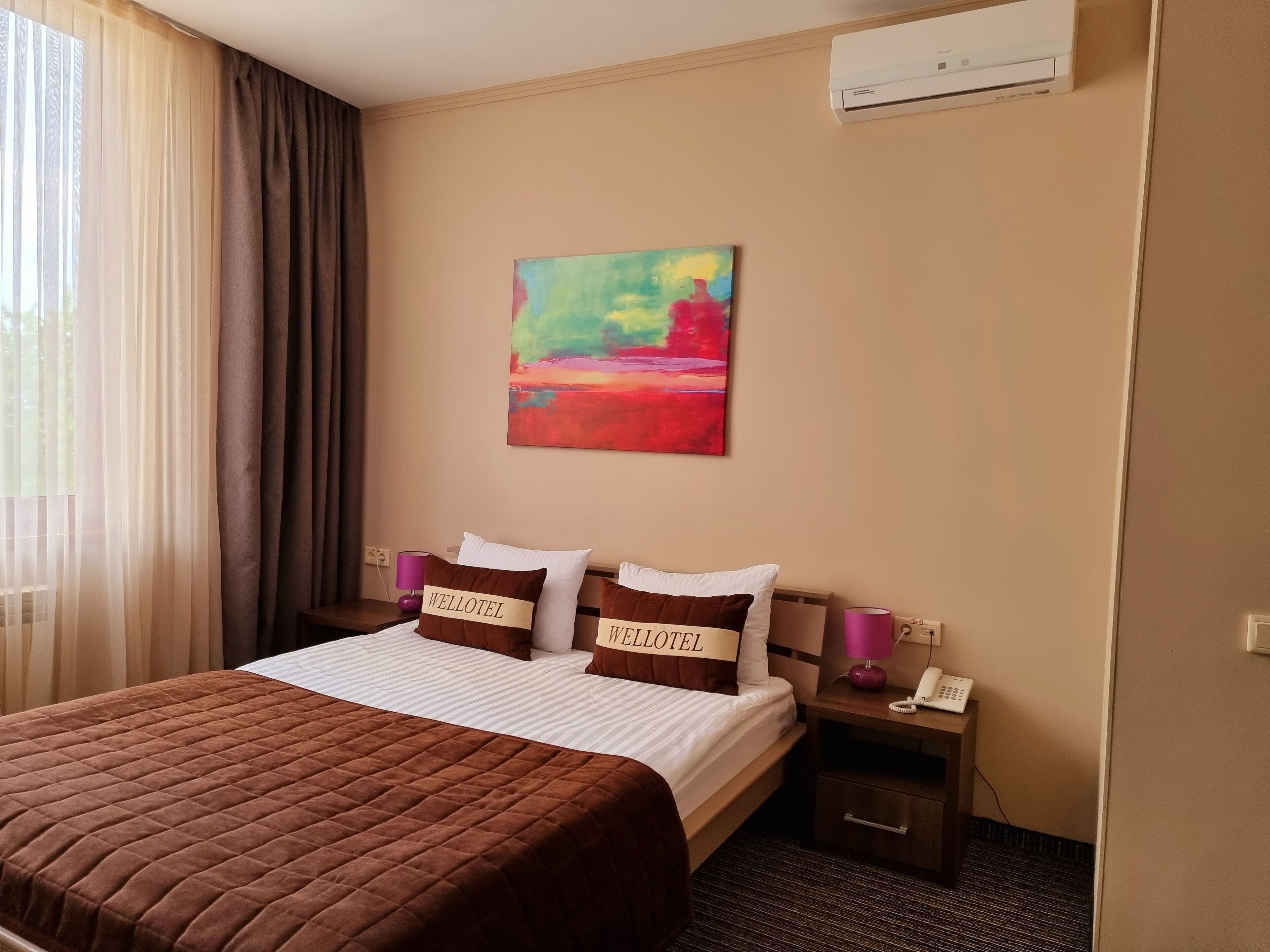 A hotel room with a bed, a painting, an air conditioner, a nightstand with a lamp, and a phone.