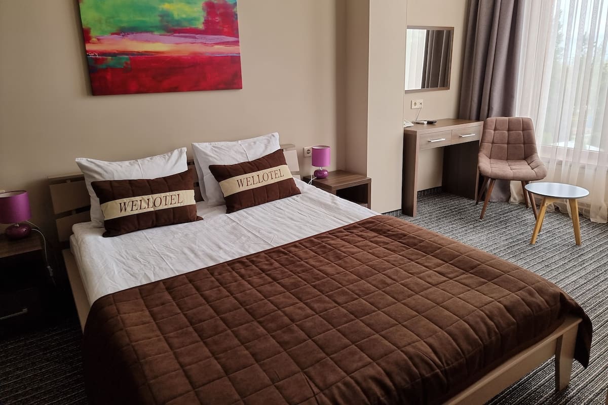 Ein Hotelzimmer mit einem Bett, zwei Kissen mit dem Schriftzug 'WELLOTEL', einem Schreibtisch, einem Stuhl, einem kleinen Tisch, einer Lampe und einem Bild an der Wand.