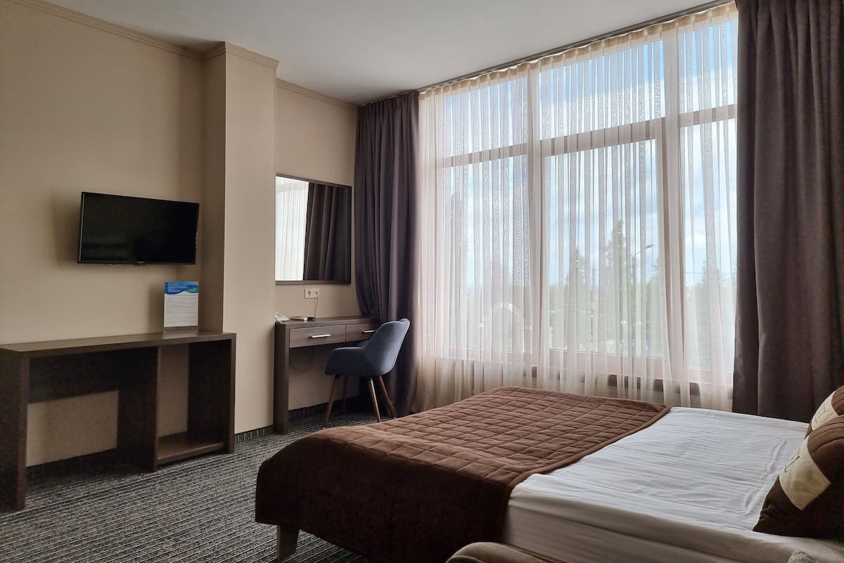 Ein Hotelzimmer mit einem Bett, einem Schreibtisch, einem Sessel, einem Fernseher und großen Fenstern mit Vorhängen.