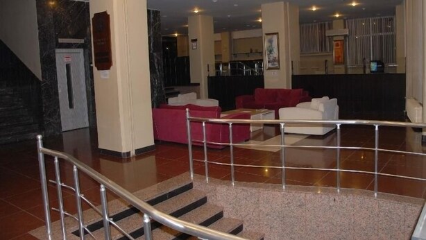 Un vestíbulo de hotel con una recepción de mármol, una escalera con barandillas metálicas y una zona de estar con sofás rojos y blancos.