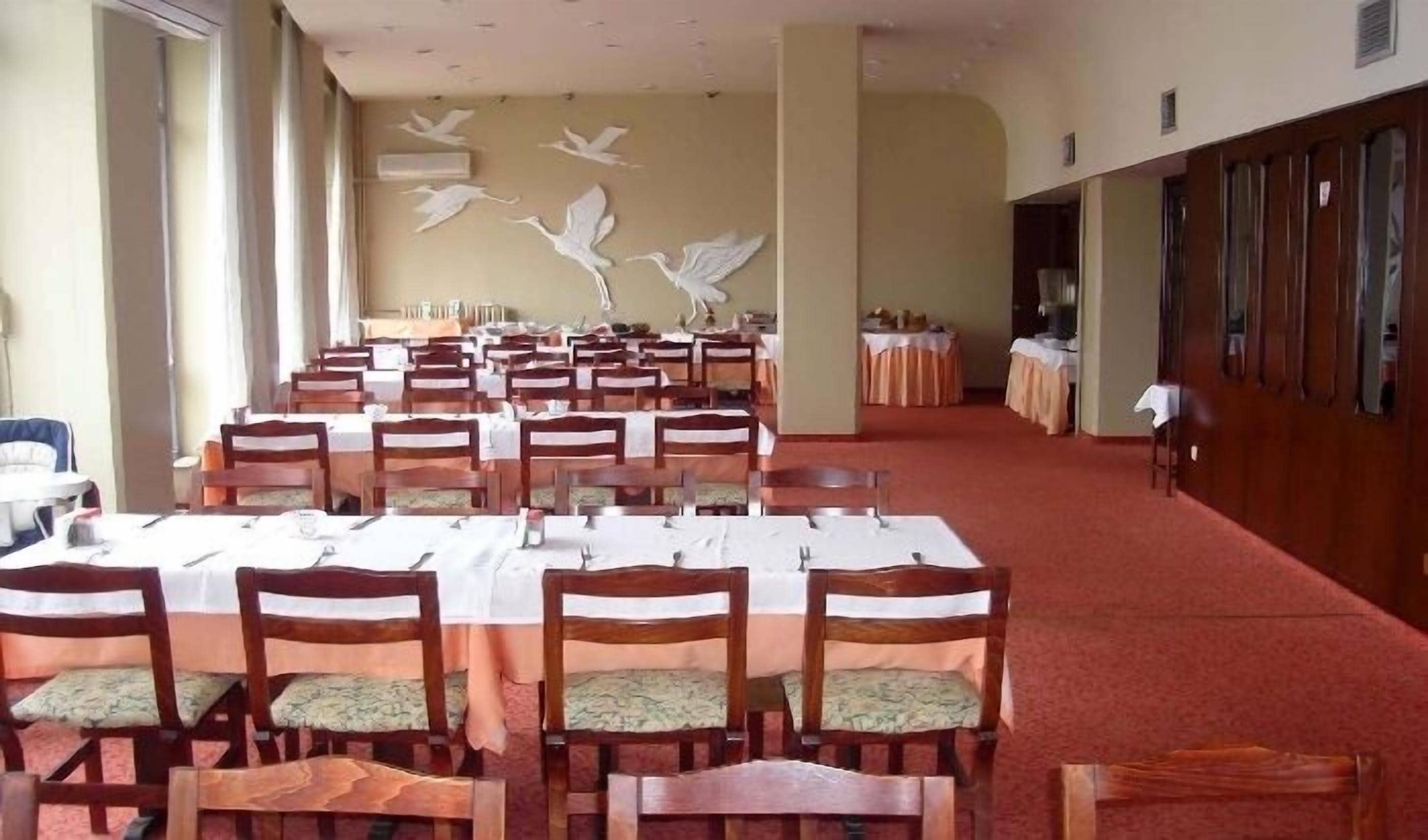 Un comedor con varias mesas y sillas, un muro decorativo con pájaros volando, y una alfombra roja.