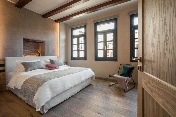 A bedroom with a large bed, wooden beams, a stone fireplace, and two windows. - J&G Suites (Chania)