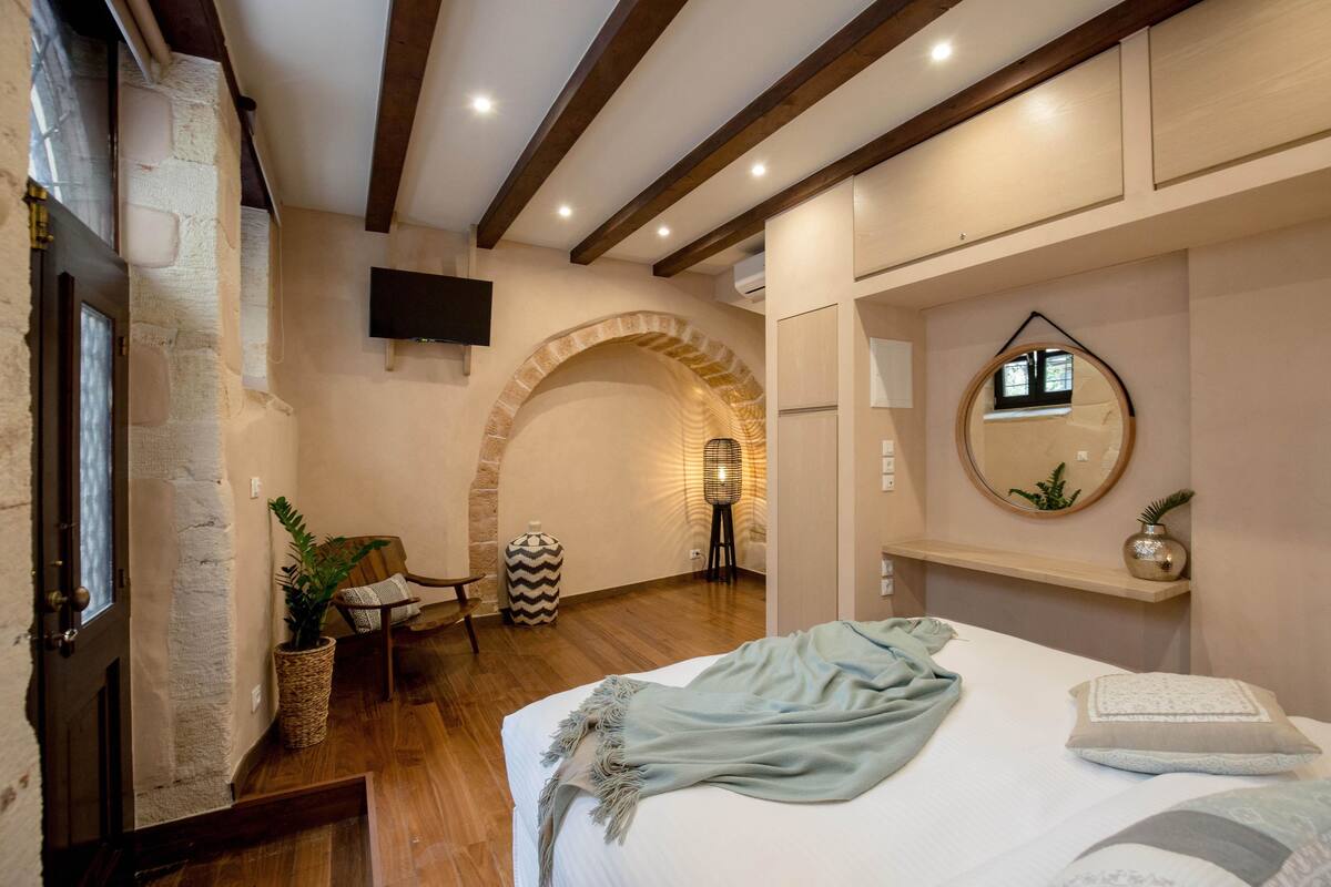 A bedroom with a bed, a wooden floor, a stone wall, a TV, a mirror, and a small table with a chair.