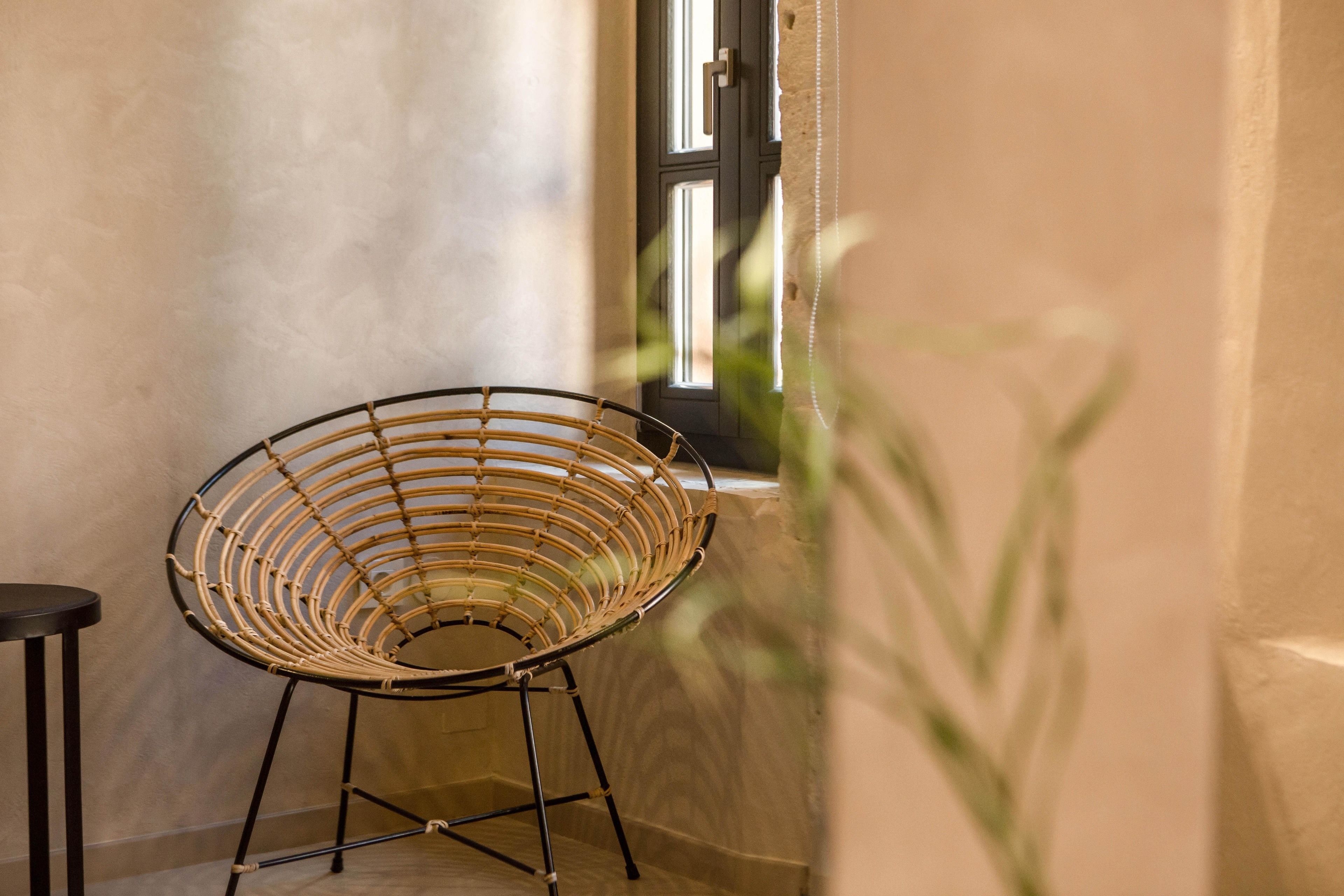 A wicker chair with a metal frame in a corner of a room with a textured wall and a window.