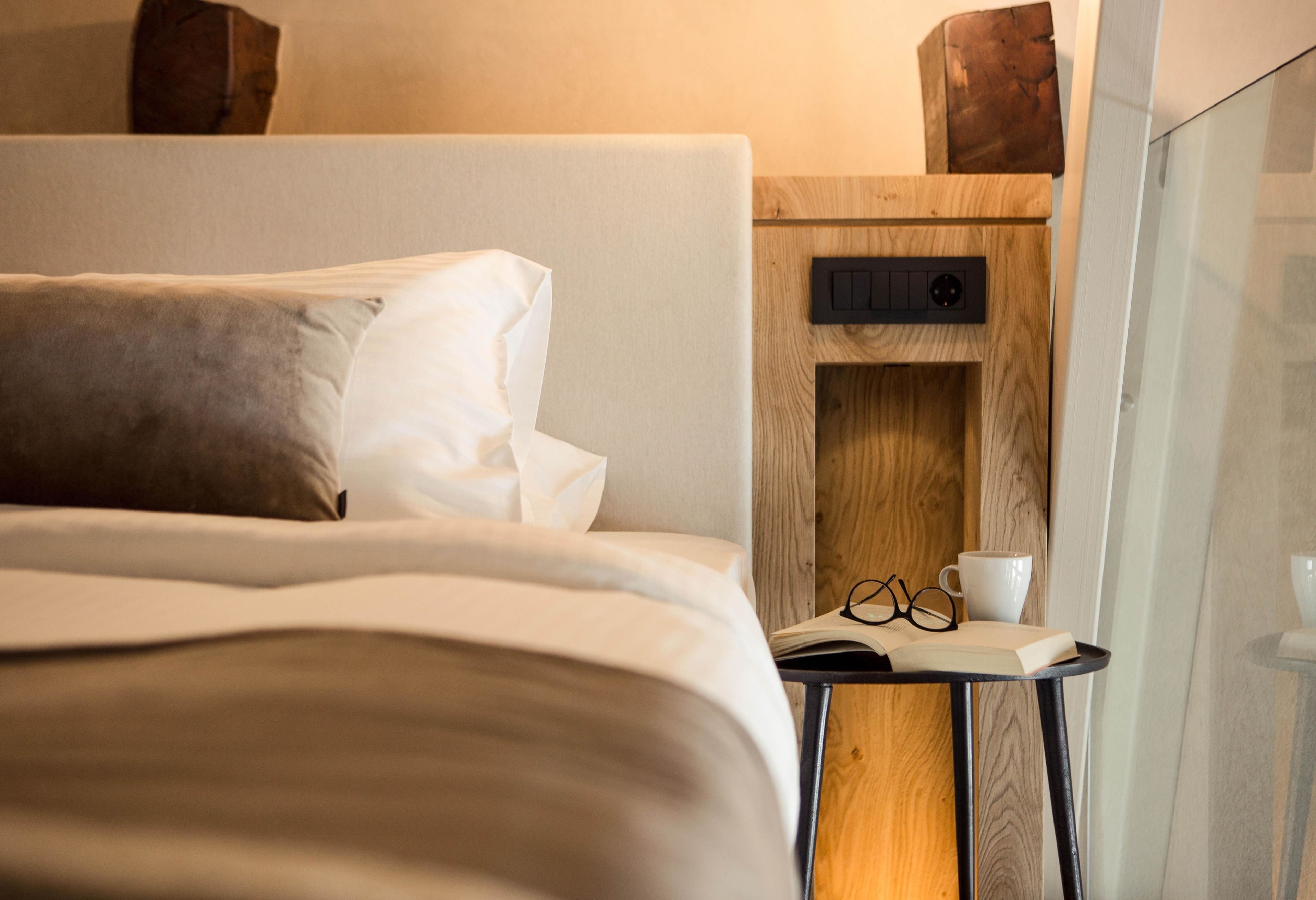 A neatly made bed with pillows, a bedside table with a book, glasses, and a cup.