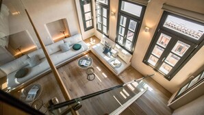 Une pièce aux hauts plafonds, dotée d’un plancher en bois, d’un canapé, d’une petite table et d’une fenêtre donnant sur des immeubles.