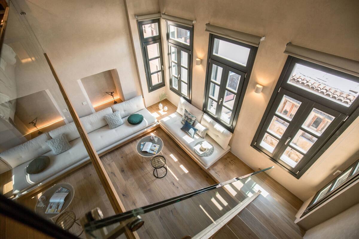 A high-ceilinged room with a wooden floor, a sofa, a small table, and a window with a view of buildings.