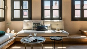 Un salon chaleureux avec un canapé, une table basse ronde et une petite table d’appoint. De grandes fenêtres aux montures sombres laissent entrer la lumière naturelle.