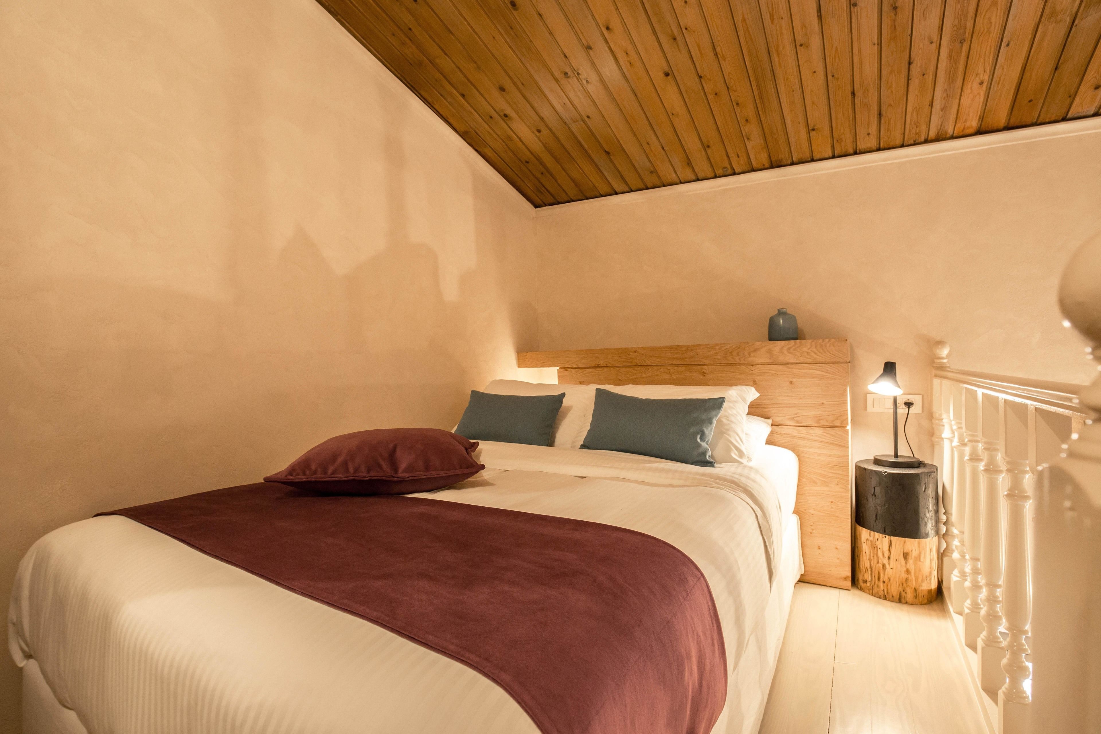Een slaapkamer met een houten plafond, een bed met wit en bordeauxrood beddengoed, en een nachtkastje met een lamp.