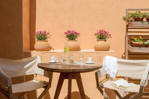 Une table ronde en bois, entourée de chaises, avec un livre et un verre contenant une boisson. Sur une étagère derrière la table, on aperçoit des plantes en pot ornées de fleurs roses.