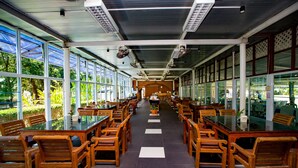 A long, open-plan dining area with wooden tables and chairs, large windows, and ceiling fans. - The Princess Beach Resort (Klaeng)