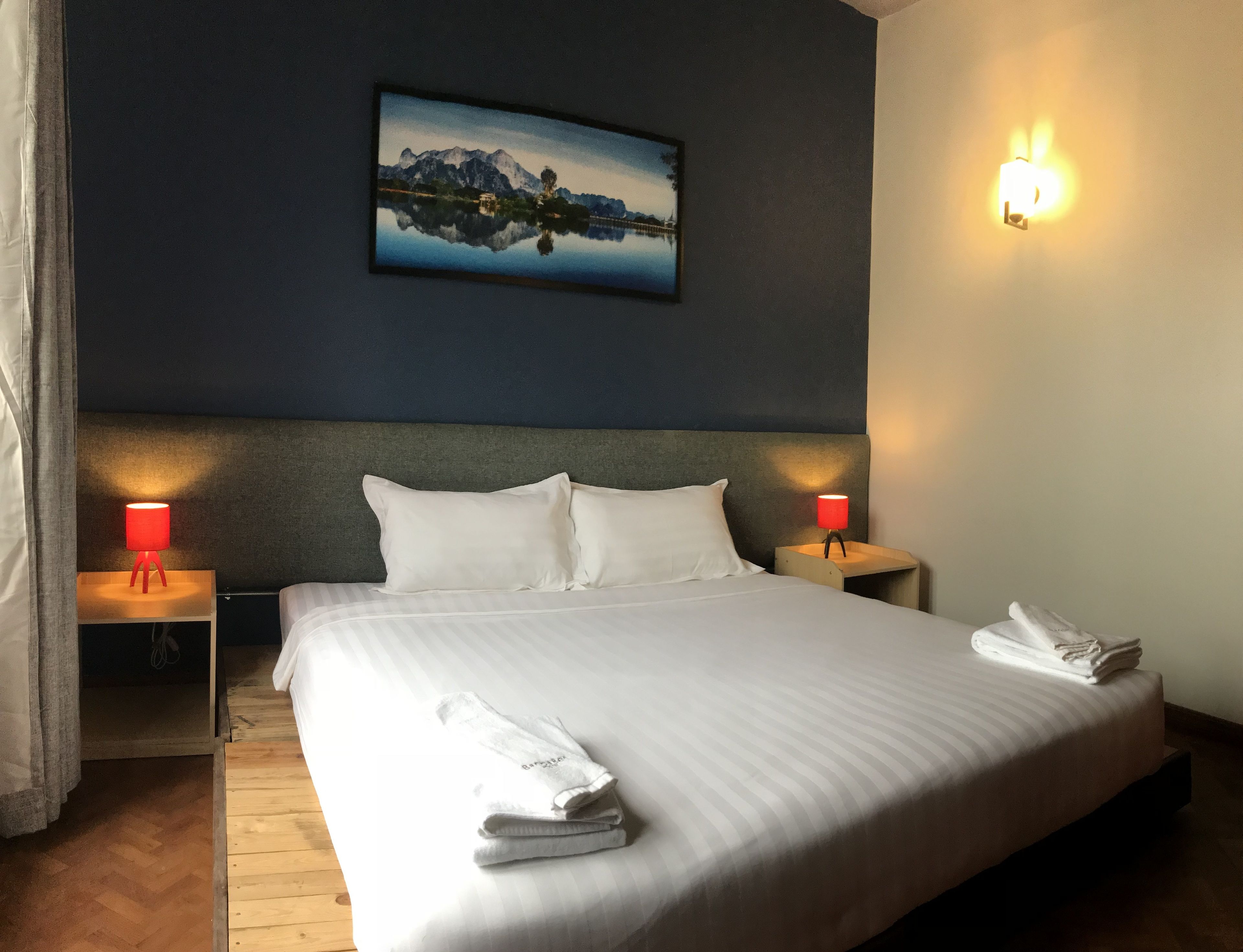 A neatly made bed with white linens and two pillows, a headboard, a wall-mounted painting of a mountain landscape, and two bedside lamps with red shades.