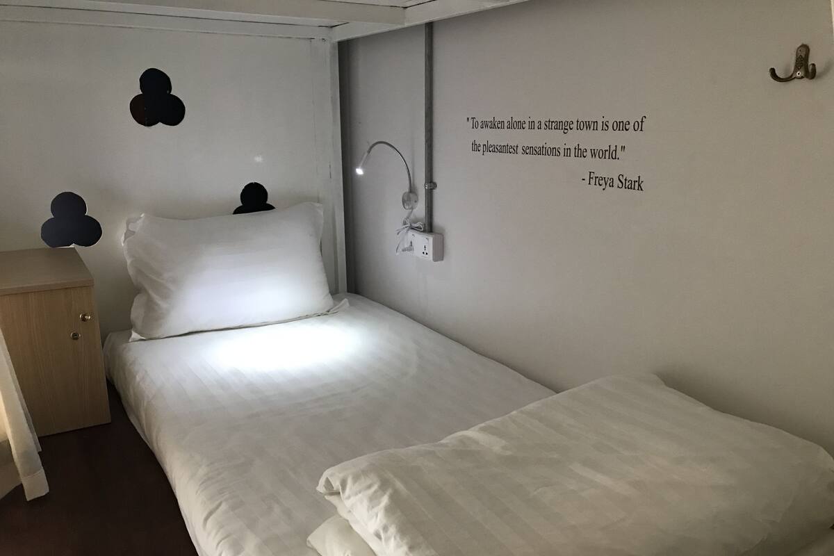 Een net opgemaakt bed met witte lakens in een kamer met een strak, minimalistisch design. Er is een wandlamp te zien en een citaat van Freya Stark staat op de muur.