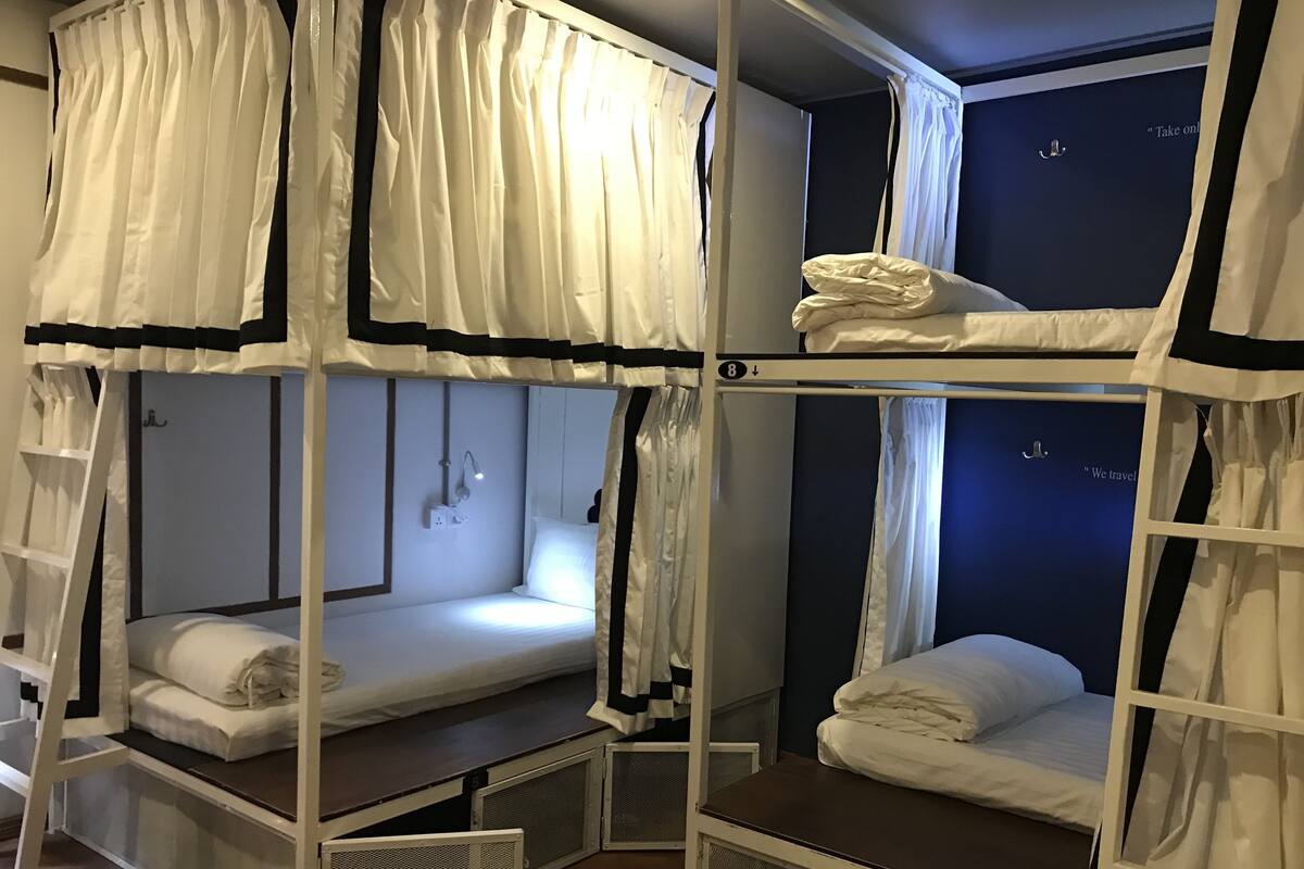 Een slaapkamer met een stapelbed, witte beddengoed, een klein bureau en een raam met gordijnen.