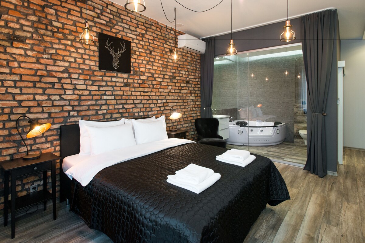 Una habitación de hotel moderna con pared de ladrillo, una cama grande con ropa blanca, cabecero negro, una mesita de noche pequeña con lámpara, una silla, un jacuzzi con paredes de vidrio y un ventanal con cortinas.