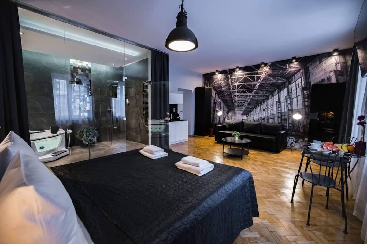 A modern hotel room with a large bed, a sitting area with a sofa and coffee table, a glass-enclosed bathroom, and a dining area with a table and chairs.