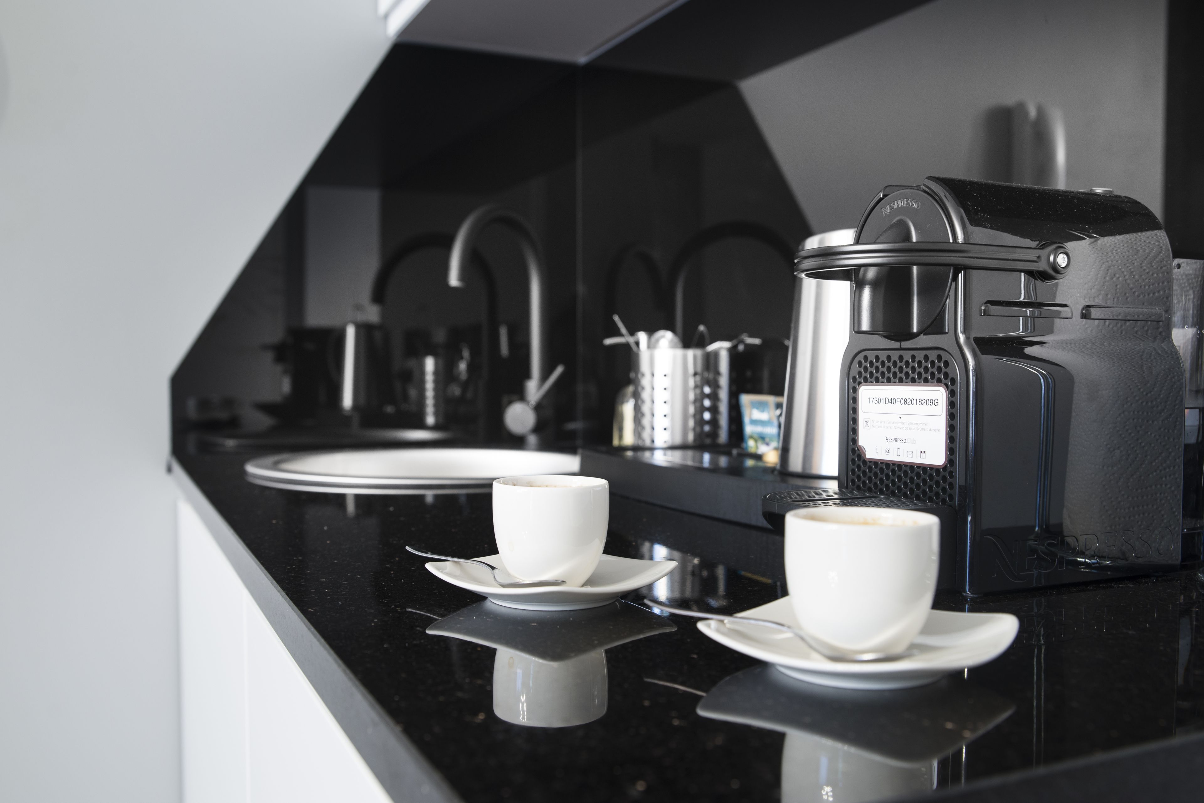 Una cocina moderna con una encimera negra, dos tazas blancas sobre platillos, una cafetera y un microondas.