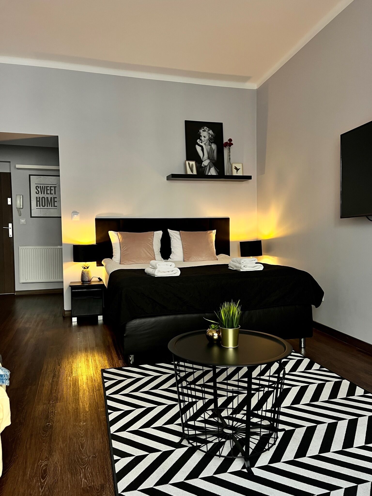 Une chambre d’hôtel moderne avec un lit soigneusement fait, une table de chevet avec une lampe, une petite table ronde agrémentée d’une plante et un tapis aux motifs noir et blanc.