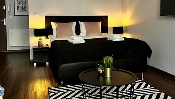 Une chambre d’hôtel moderne avec un lit soigneusement fait, une table de chevet avec une lampe, une petite table ronde agrémentée d’une plante et un tapis aux motifs noir et blanc.