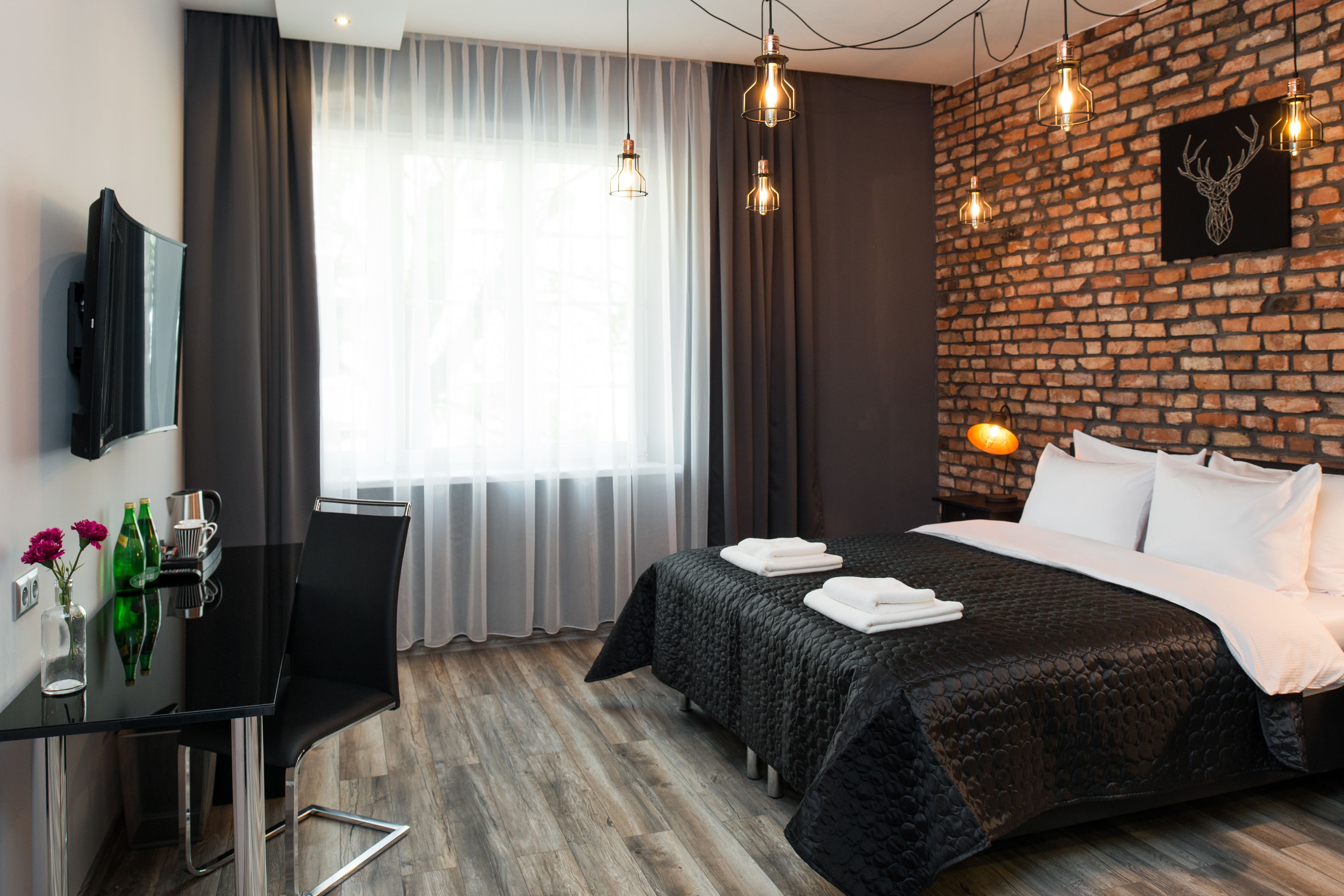 Ein modernes Hotelzimmer mit Backsteinwand, einem großen Bett mit schwarzer Bettdecke, einem Schreibtisch mit Stuhl und einem Flachbildfernseher.