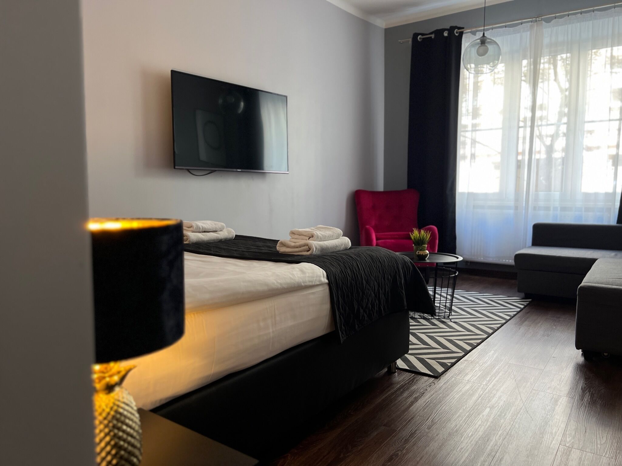A modern hotel room with a bed, a red chair, a small table with a plant, and a TV on the wall.