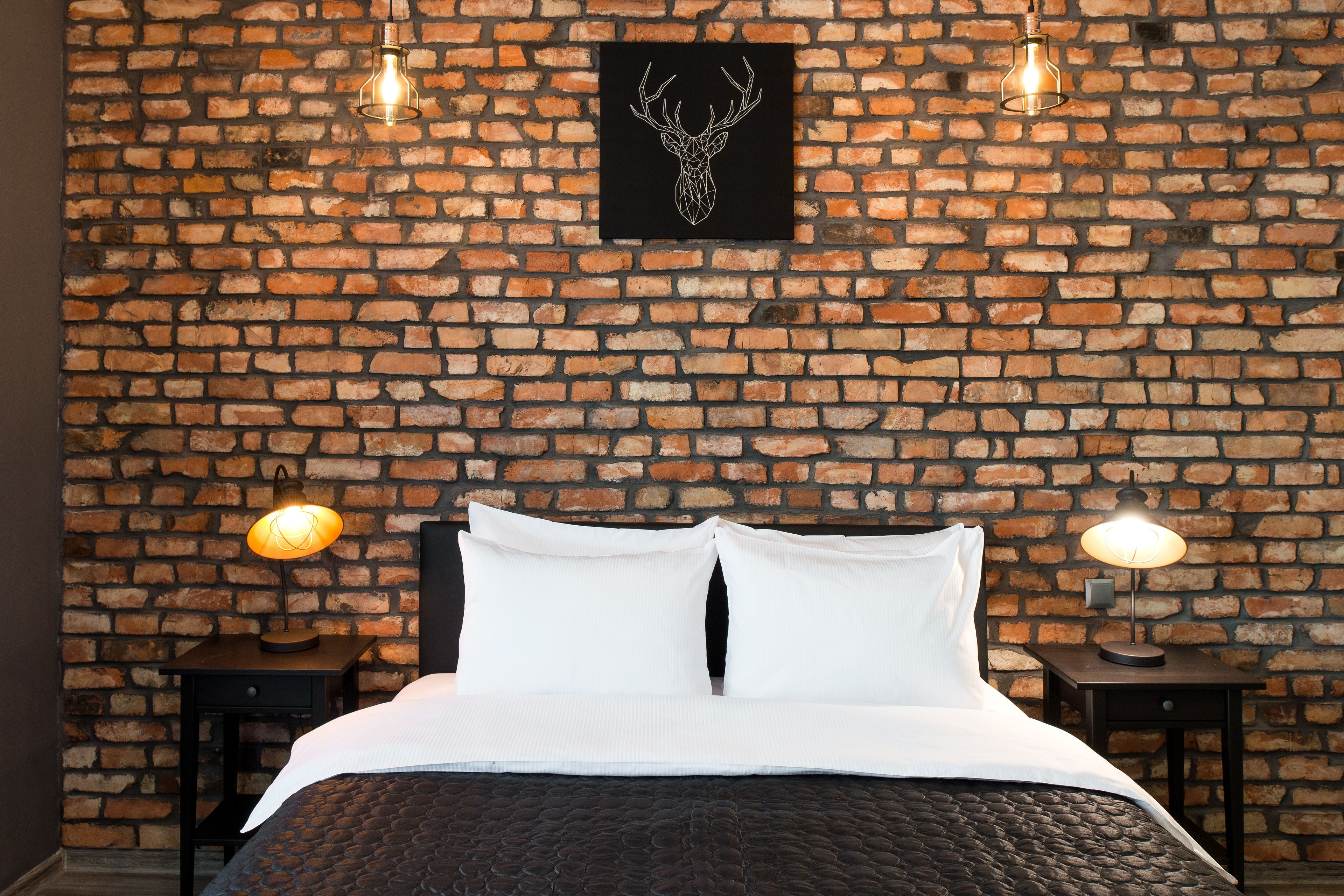 A room with a brick wall, a bed with white bedding, two bedside tables with lamps, and a framed deer head artwork on the wall.