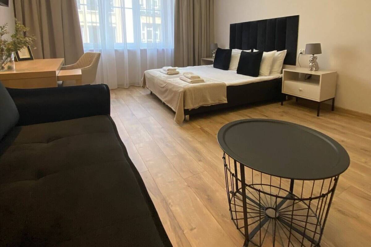 A modern hotel room with a bed, a sofa, a round dining table, and a TV mounted on the wall.
