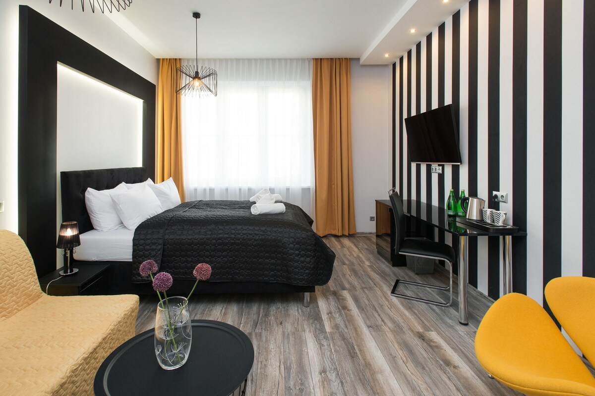 A modern hotel room with a large bed, a desk with a flat-screen TV, a sofa, and a coffee table with flowers.