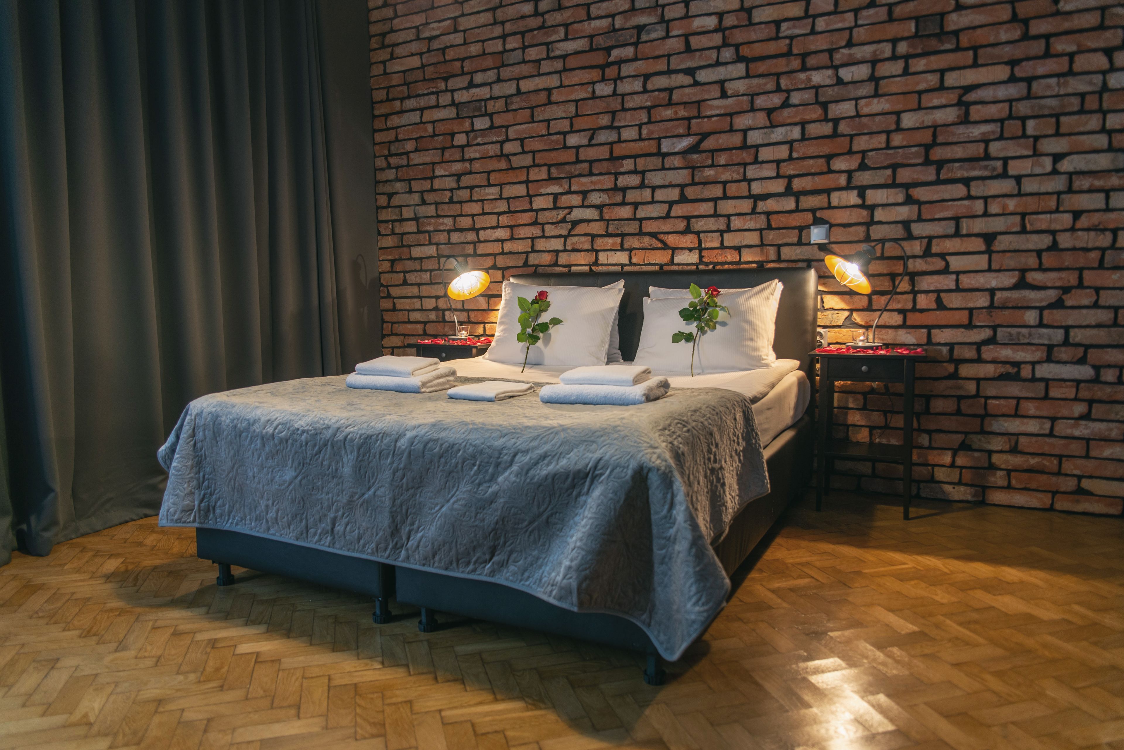 A bedroom with a brick wall, a bed with a grey blanket, two bedside lamps, and a wooden herringbone floor.