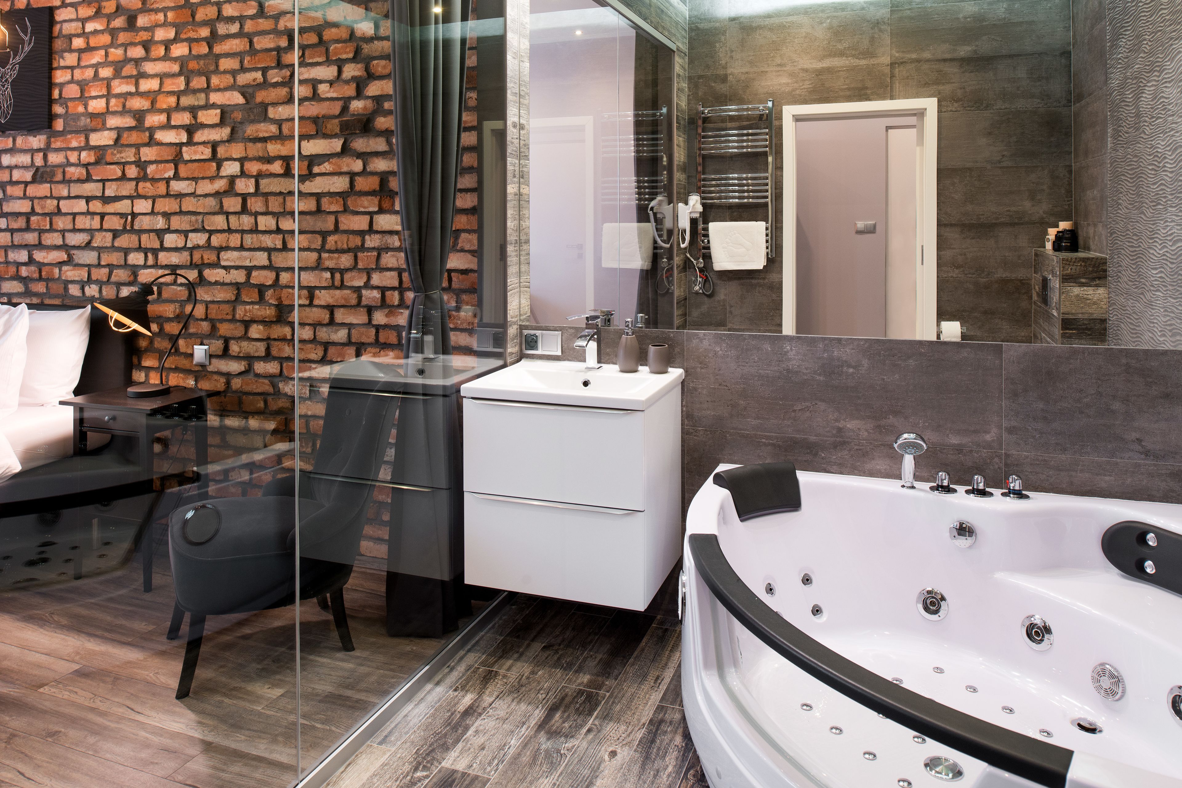 A modern bathroom with a brick wall, a white sink, a glass shower enclosure, a black chair, and a large bathtub.