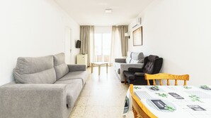 A modern living room with a grey sofa, a wooden dining table with a patterned tablecloth, and a black armchair. - Apartamentos Maja (Benidorm)