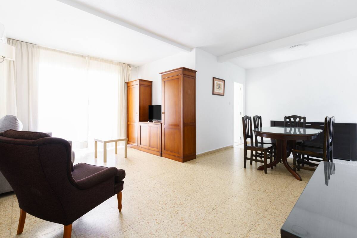 A spacious living area with a dark brown armchair, a wooden dining table with chairs, and a cabinet with a flat-screen TV.