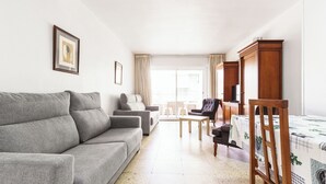 A living room with a grey sofa, a wooden chair, a small table, and a bed with a patterned bedspread. - Apartamentos Maja (Benidorm)