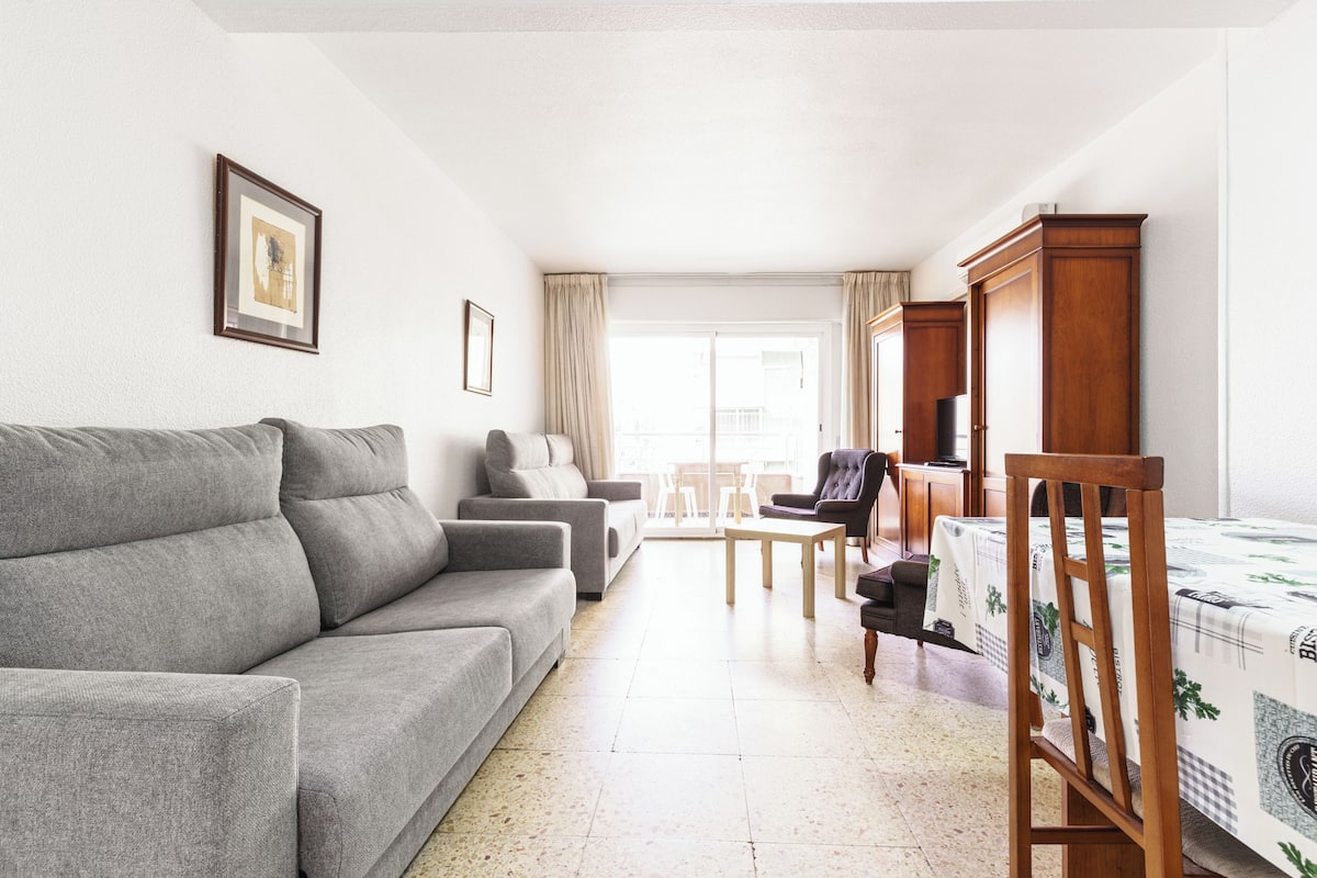 A living room with a grey sofa, a wooden chair, a small table, and a bed with a patterned bedspread.